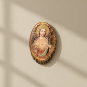 Oval-shaped, rustic wall plaque of the Sacred Heart of Jesus on a beige wall in midday lighting.