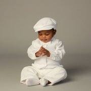 Baby in a white baptism outfit with a cap sitting on a gray background