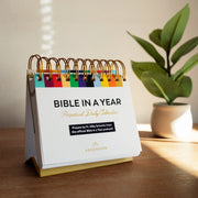 A colorful desk calendar with the text 'Bible in a Year Perpetual Daily Calendar' rests on a sunlit table with a potted plant in the background.