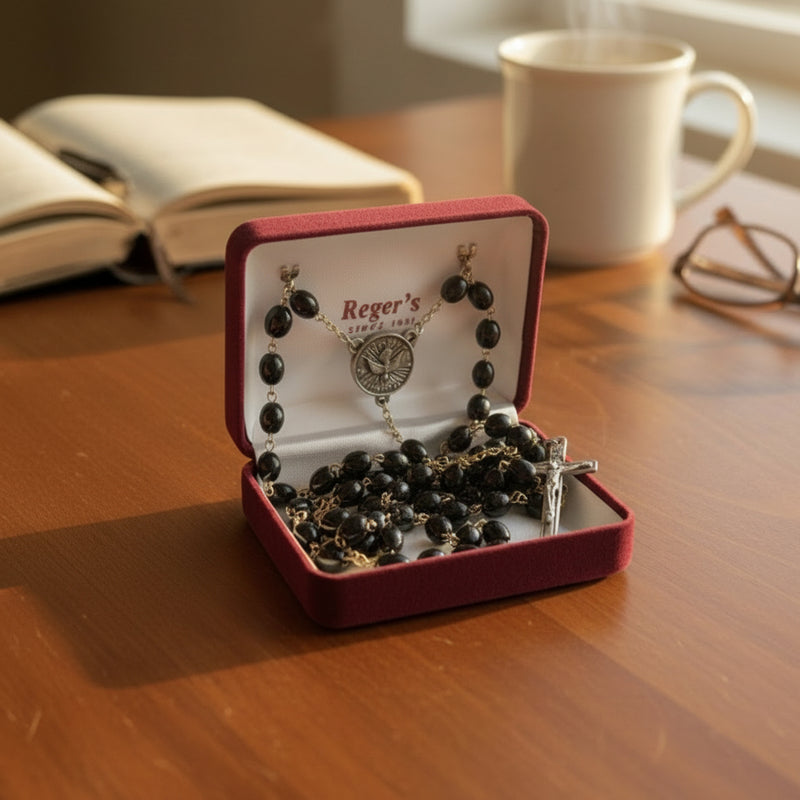 Hand holding a black Confirmation rosary in a 'Reger's' jewelry box on a wooden table.