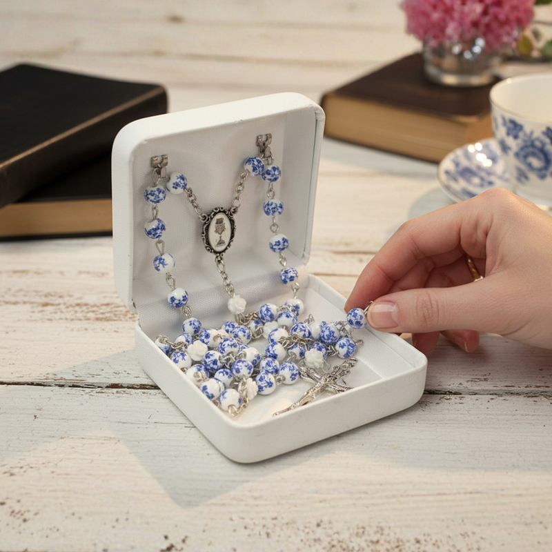 Hand holding a girl's blue and white First Communion rosary over an open jewelry box on a wooden surface.