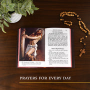 Open book with religious text and illustration on a wooden surface, accompanied by a rosary and plant.