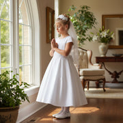Young girl in a white Communion dress standing in a sunlit room with plants and furniture.