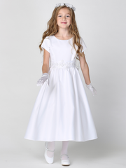 A young girl wearing a white satin First Communion dress with silver corded trim on the waist, standing against a plain background.