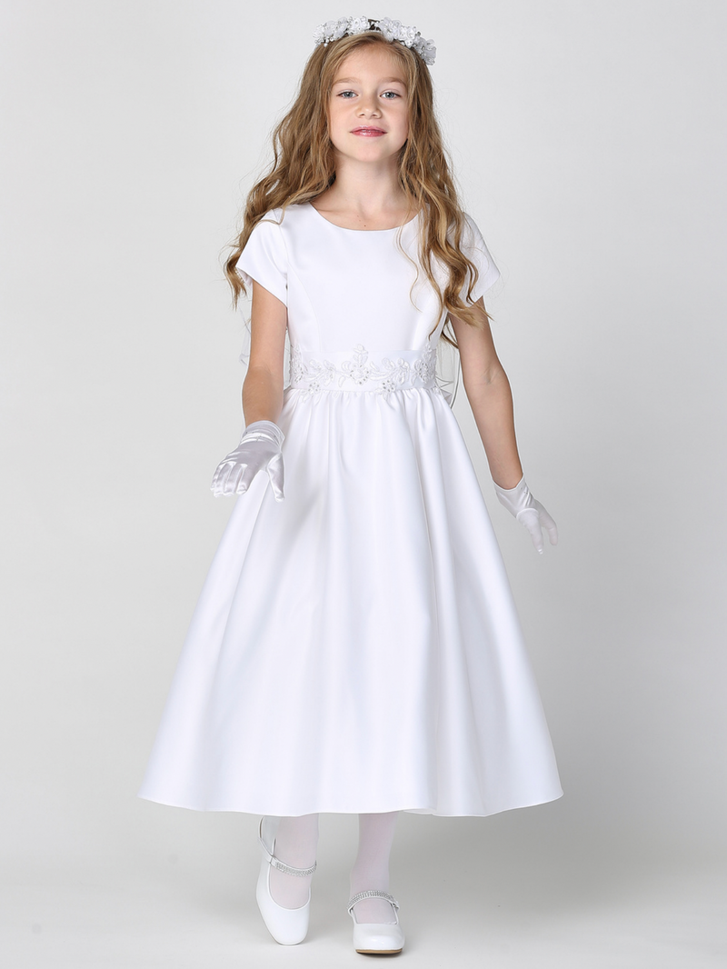 A young girl wearing a white satin First Communion dress with silver corded trim on the waist, standing against a plain background.