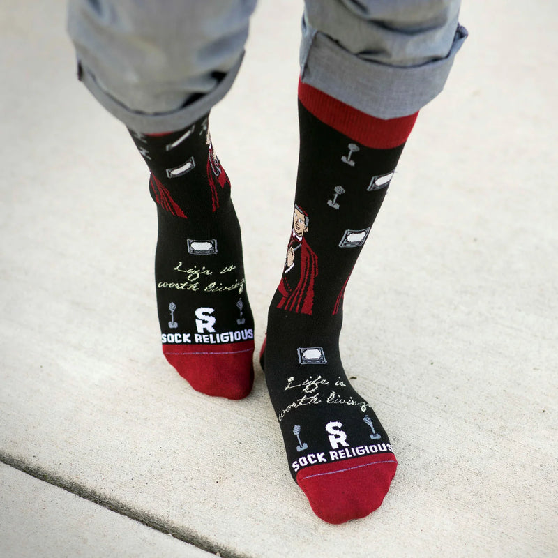 Person wearing black Fulton Sheen socks with red accents on a light gray pavement.