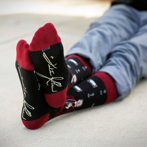 Person wearing black Fulton Sheen socks with red accents on a light gray pavement.