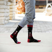 Person wearing black Fulton Sheen socks with red accents on a light gray pavement.