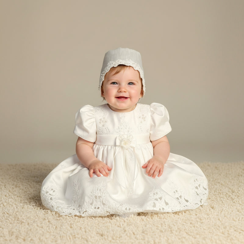 Baby in a white baptismal dress and bonnet sitting on a beige carpet.