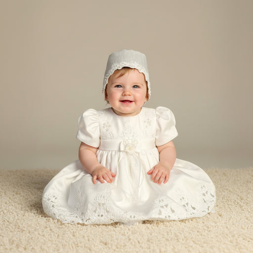 Baby in a white baptismal dress and bonnet sitting on a beige carpet.