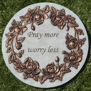 Decorative stone plaque with floral designs and text 'Pray more worry less' on grass.