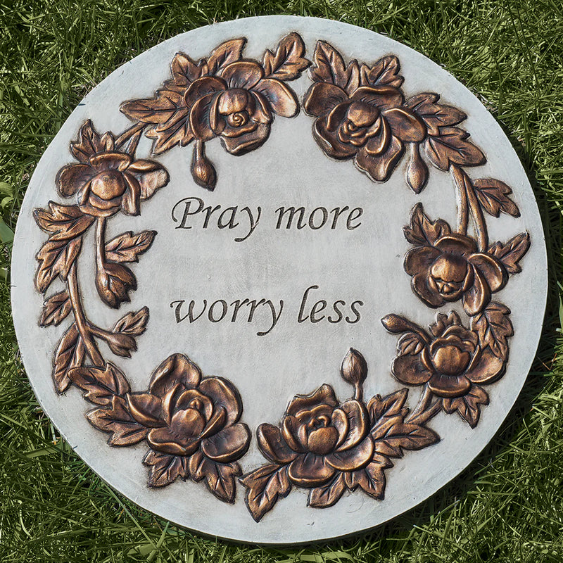 Decorative stone plaque with floral designs and text 'Pray more worry less' on grass.
