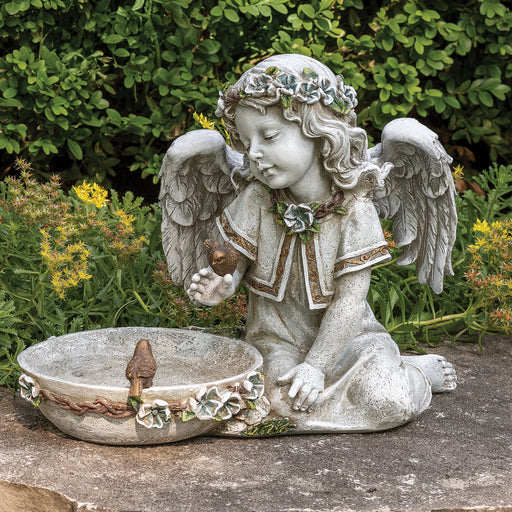 Statue of an angel with wings holding a shell, sitting by a bowl in a garden setting.