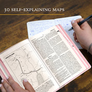 Open book with text and maps on a wooden surface, with a hand holding a pen.