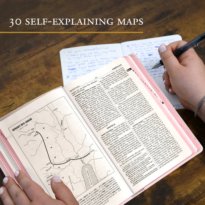 Open book with text and maps on a wooden surface, with a hand holding a pen.