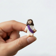 A tiny rubber Jesus with a purple sash held by someone's fingers over a gray background.
