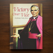 A book titled "Victory Over Vice: Archbishop Fulton Sheen" lying on a dark wooden surface. The cover features a portrait of Archbishop Fulton Sheen in clerical vestments with a bright pink cape and black cassock.