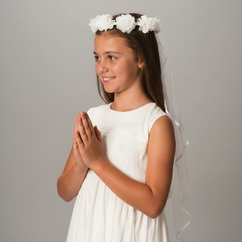 Young girl in a white Communion dress with a floral headband and veil against a gray background