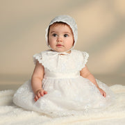 Baby in a white lace baptism dress and bonnet sitting on a soft surface with a neutral background