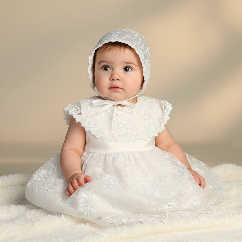 Baby in a white lace baptism dress and bonnet sitting on a soft surface with a neutral background