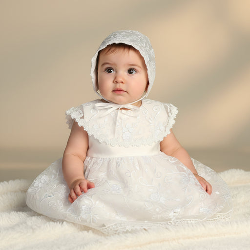 Baby in a white lace baptism dress and bonnet sitting on a soft surface with a neutral background