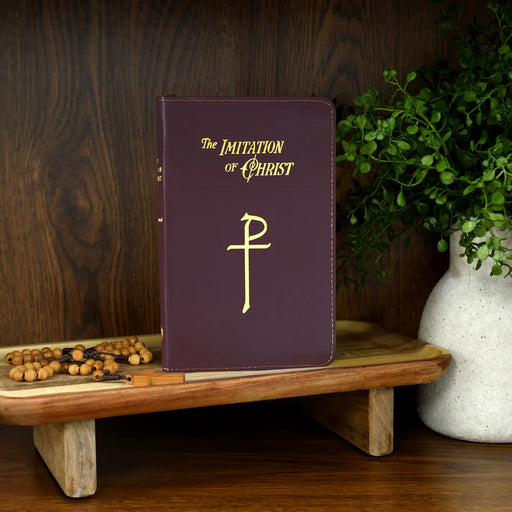 Burgundy book titled 'The Imitation of Christ' on a wooden stand with rosary and plant.