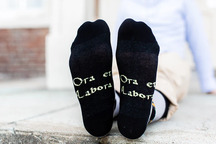 Gray socks with St. Benedict design worn by a person on a light gray pavement background
